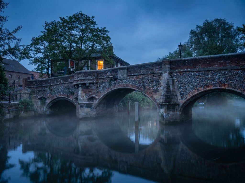 Bishop Bridge in Norwich at blue-hour dusk, three medieval flint arches reflected in the River Wensum with mist rising off the water